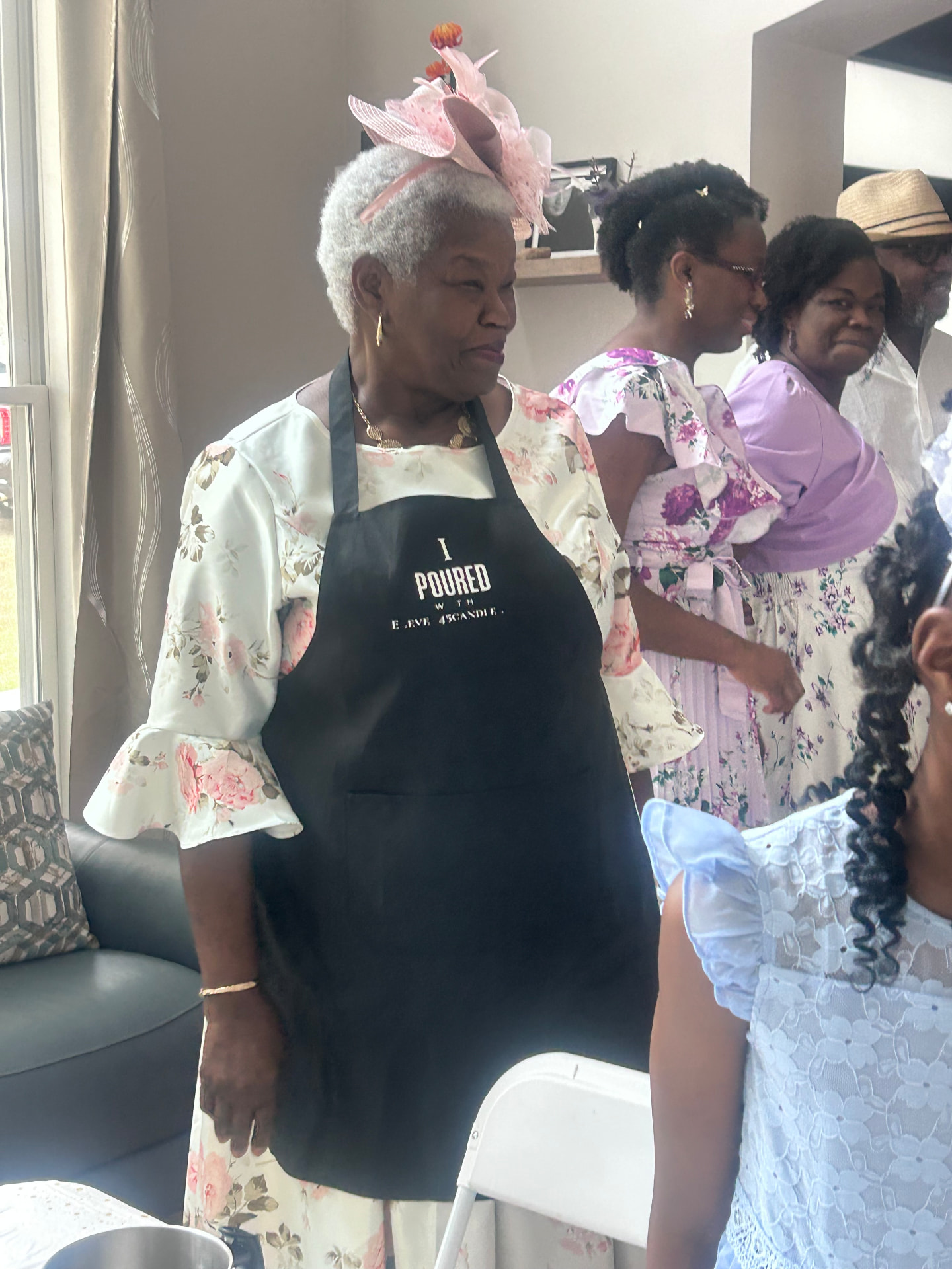 A woman in a floral dress, pink fascinator, and I Poured With Eleven45 apron among guests at a daytime gathering