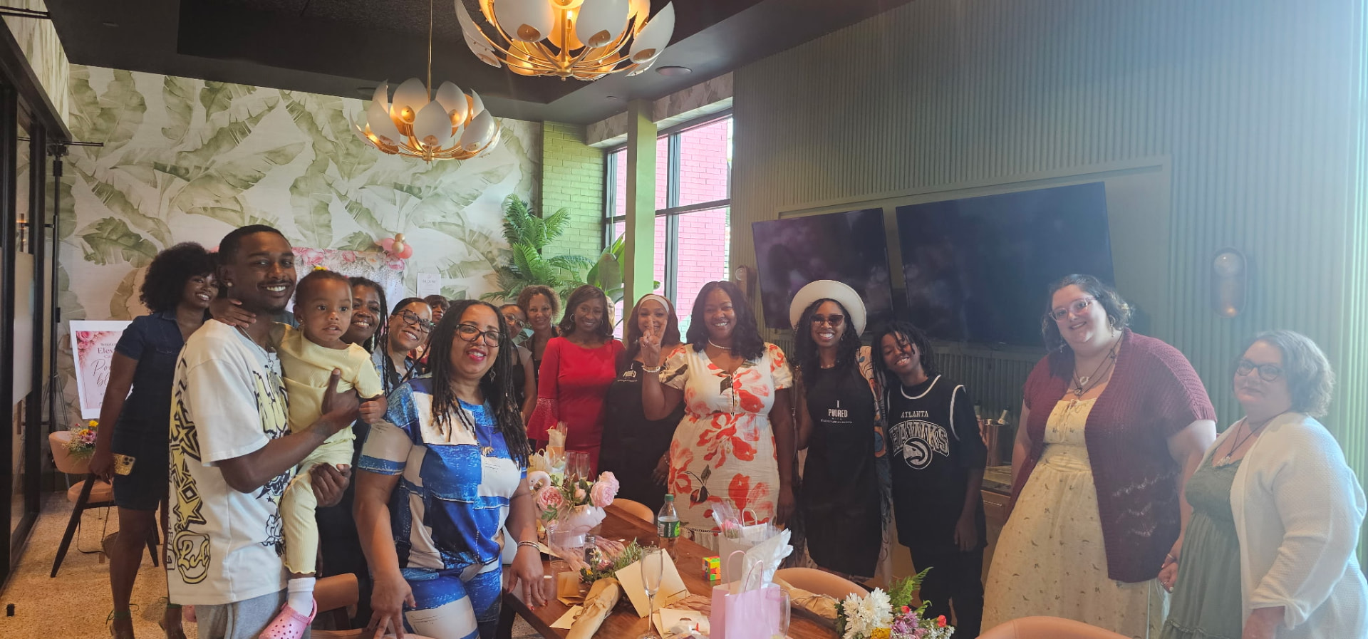 A large group of guests of all ages posing around a flower-dressed table in a room with palm-leaf wallpaper and gold chandeliers