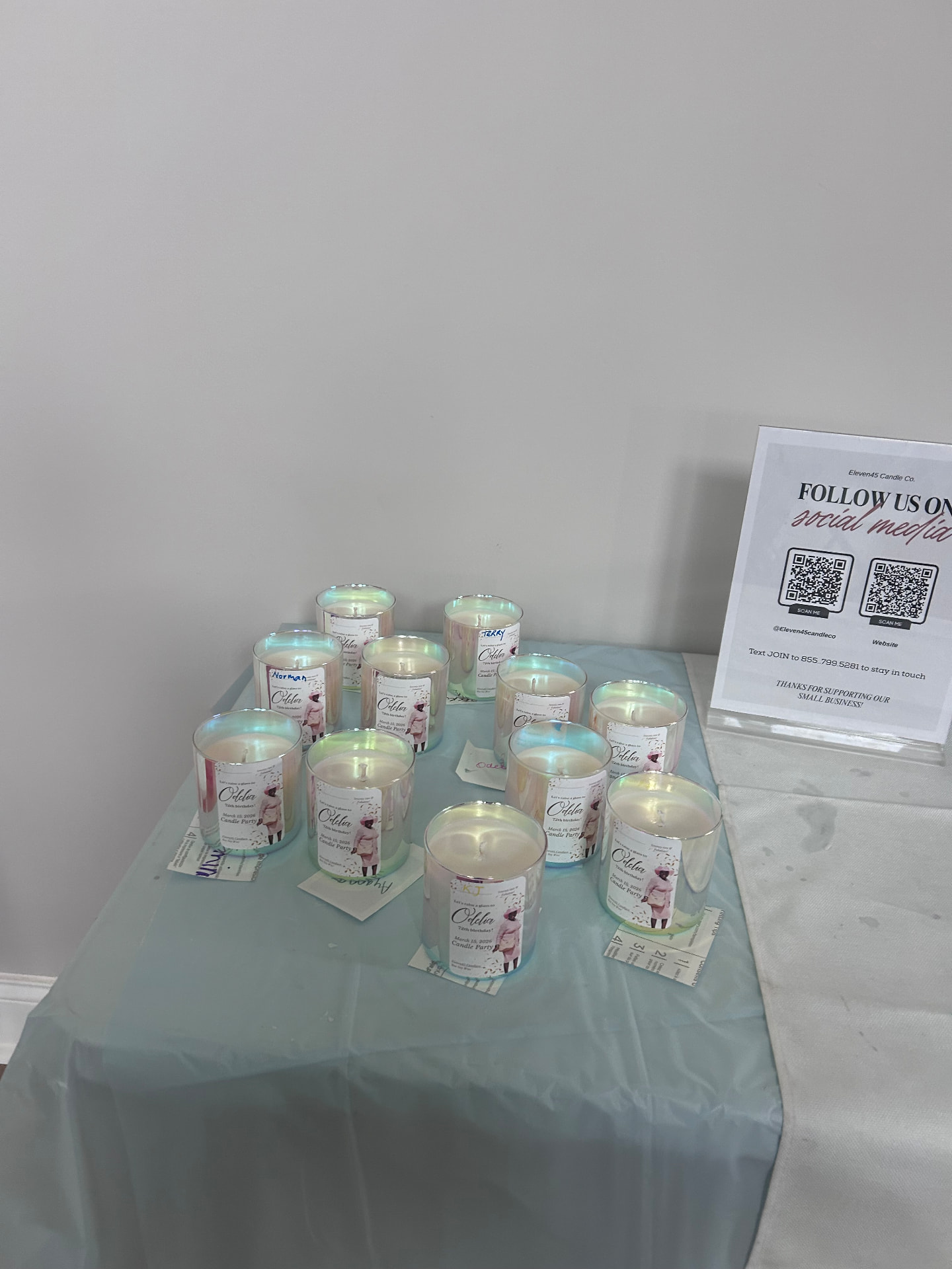 Finished candles in iridescent vessels with custom birthday party labels, displayed beside an Eleven45 Follow Us sign