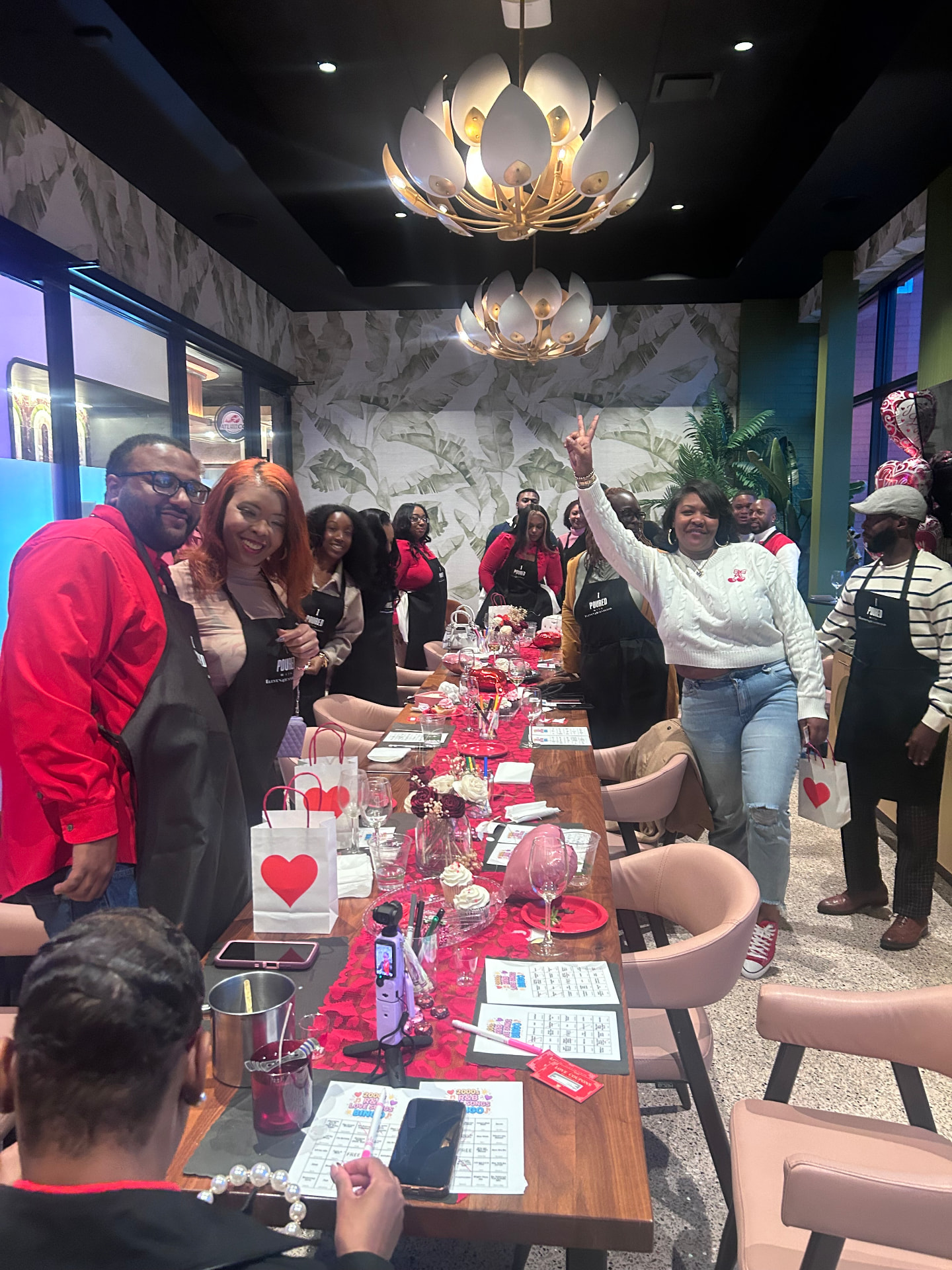 Guests in red and black I Poured With Eleven45 aprons celebrating around a long Valentine's table with heart gift bags under gold lotus chandeliers