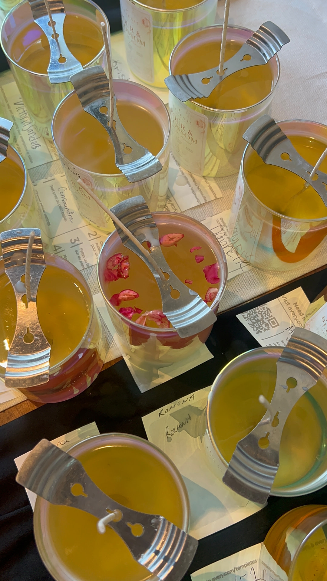 Overhead view of freshly poured candles in iridescent glass vessels with wick holders, rose petals, and handwritten name cards