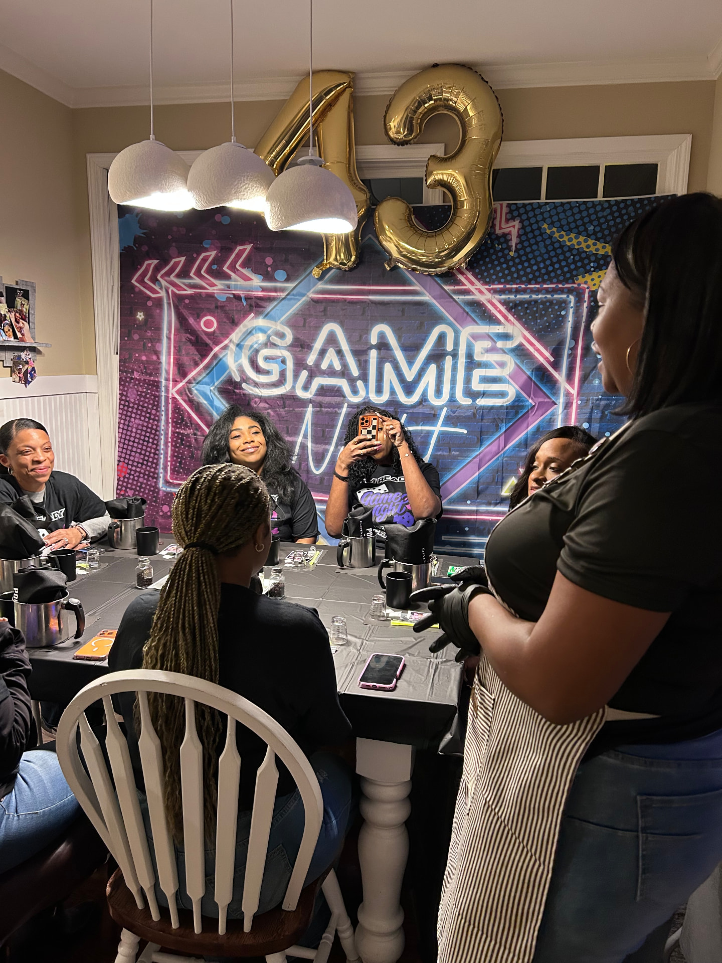 The Eleven45 instructor in a striped apron leading guests at a game night candle party under gold 43 balloons