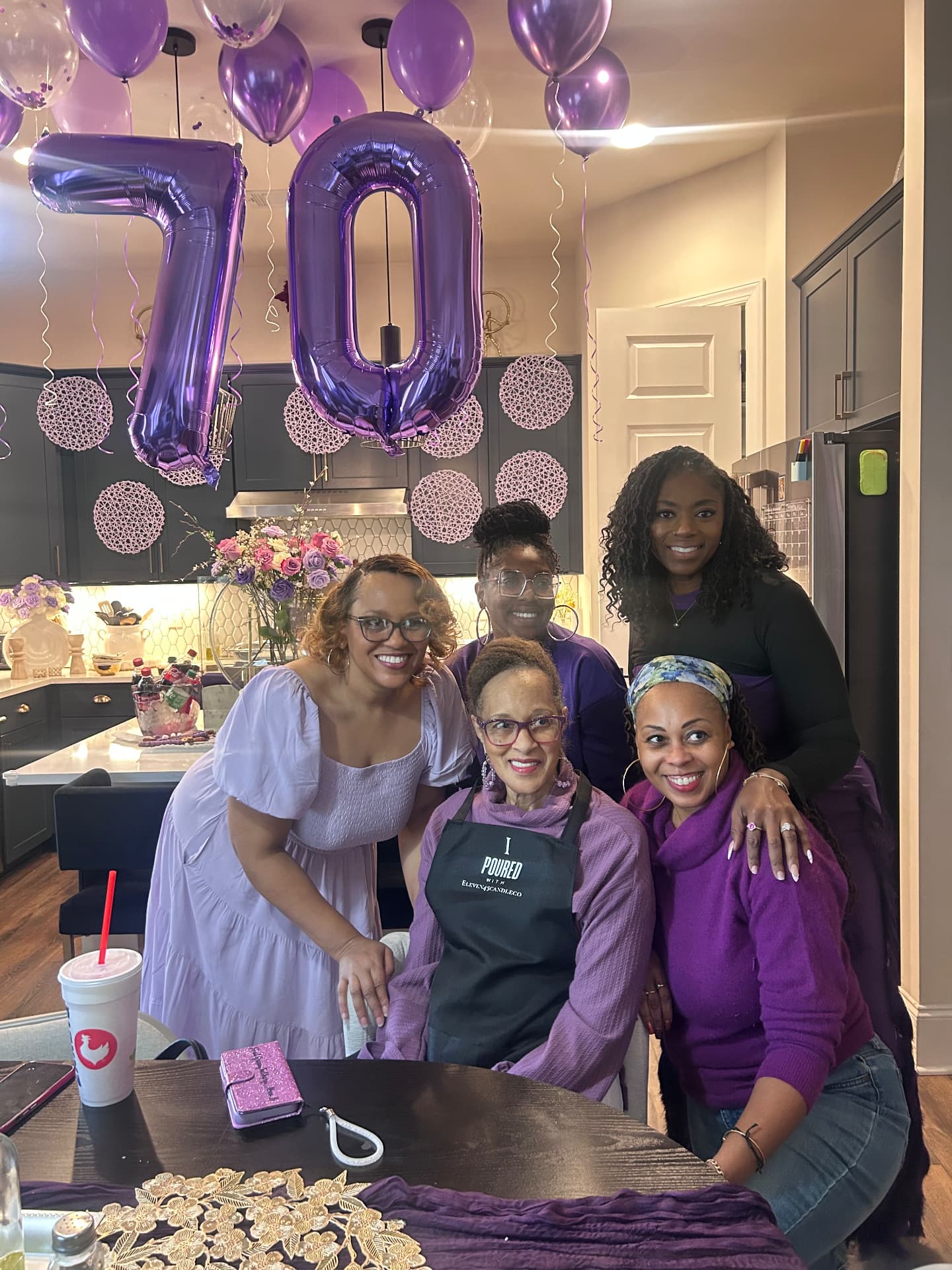 Five women in shades of purple smiling together under giant purple 70 balloons at a candle-making birthday celebration