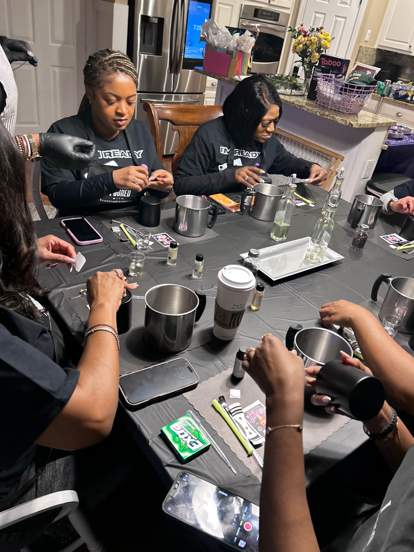 Guests in black aprons focused on preparing wicks and vessels around a kitchen table set with pouring pitchers and fragrance oils