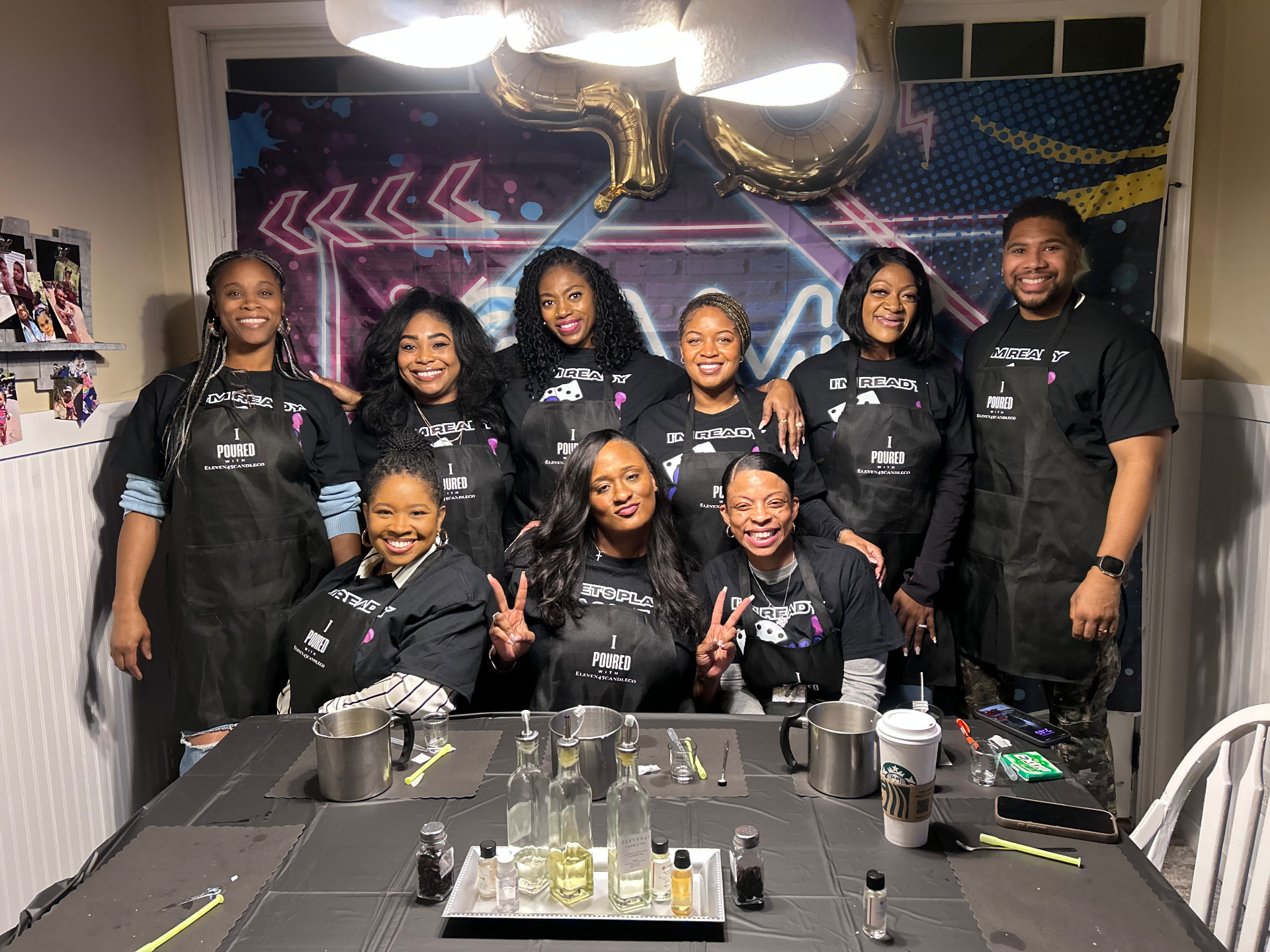 Nine smiling guests in matching black I Poured With Eleven45 aprons posing behind a candle-making table at a birthday celebration